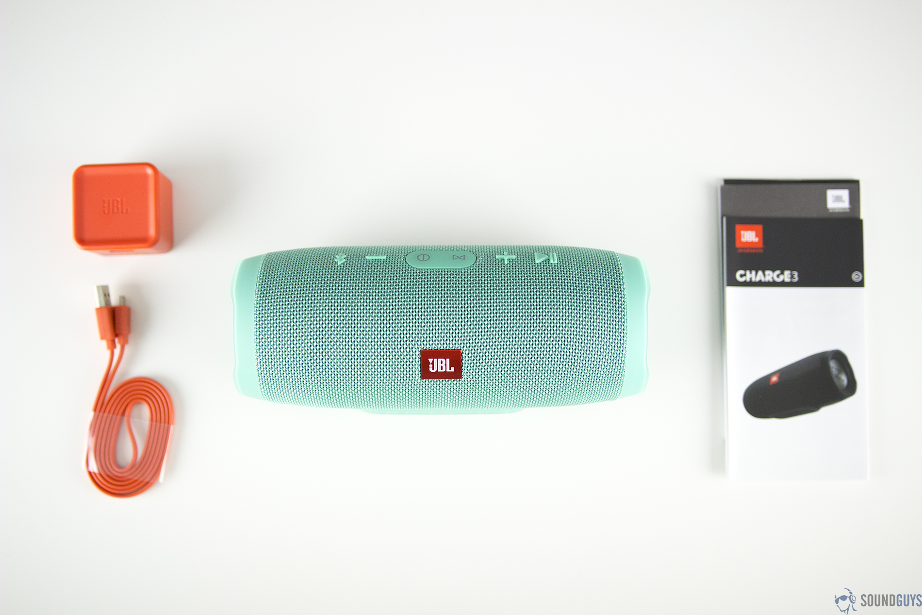 The contents of the teal JBL Charge 3 which includes an orange charging brick and cable and product information booklets on a white table.