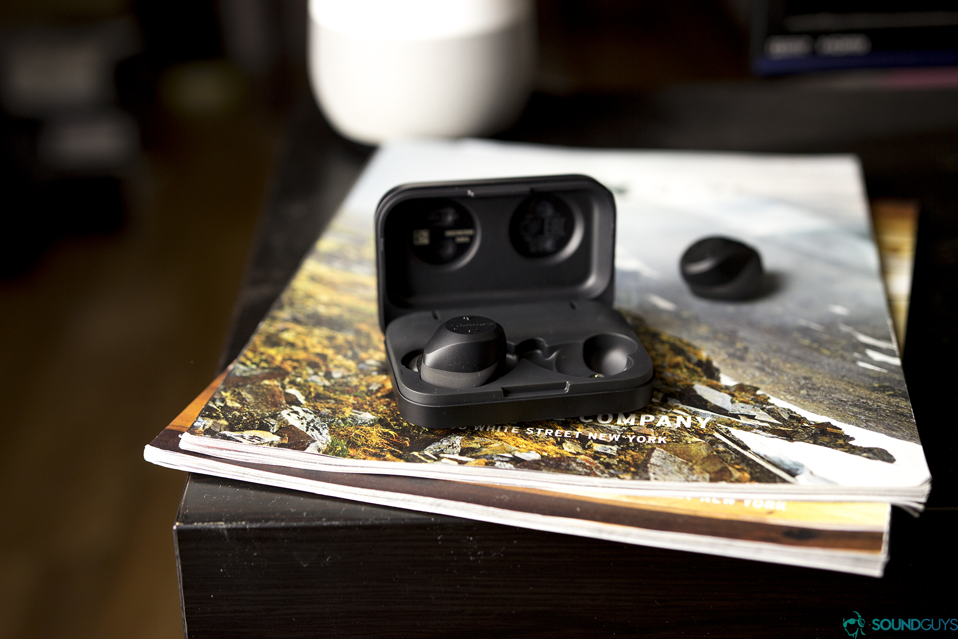 Pictured is the open charging case with one Jabra Elite Sport earbud in on top of magazines. 