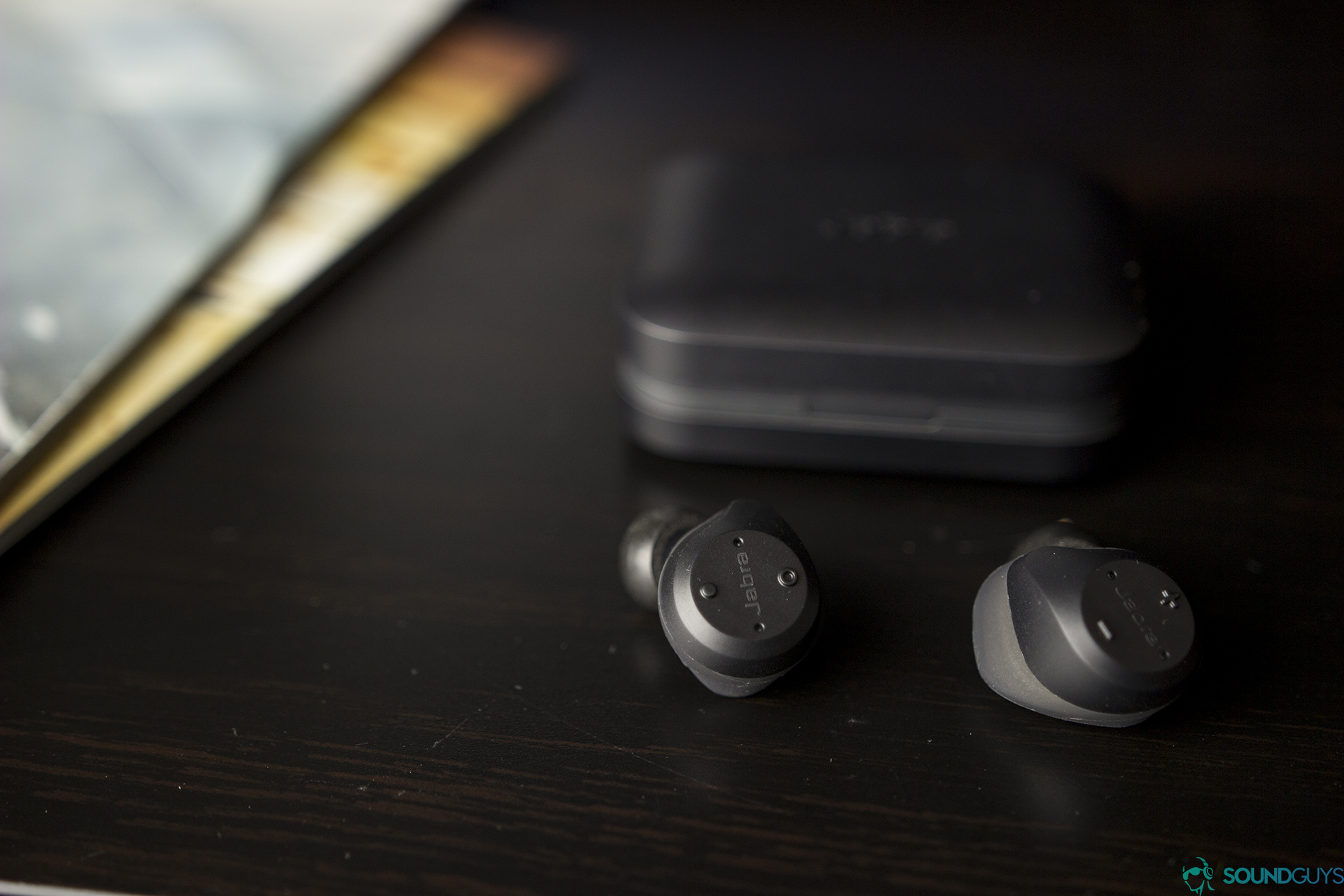 A photo of the Jabra Elite Sport true wireless earbuds on a wooden surface with the charging case in the background.