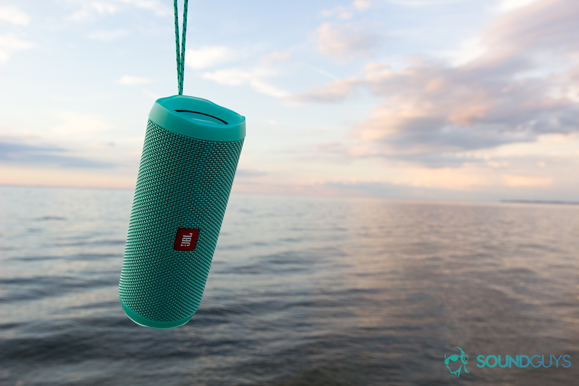 The JBL Flip 4 speaker dangling over a body of water; skies are minimally cloudy around sunset.