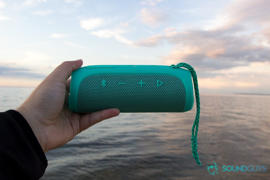 Man holding teal JBL Flip 4 in hand over the water with the playback buttons visible.