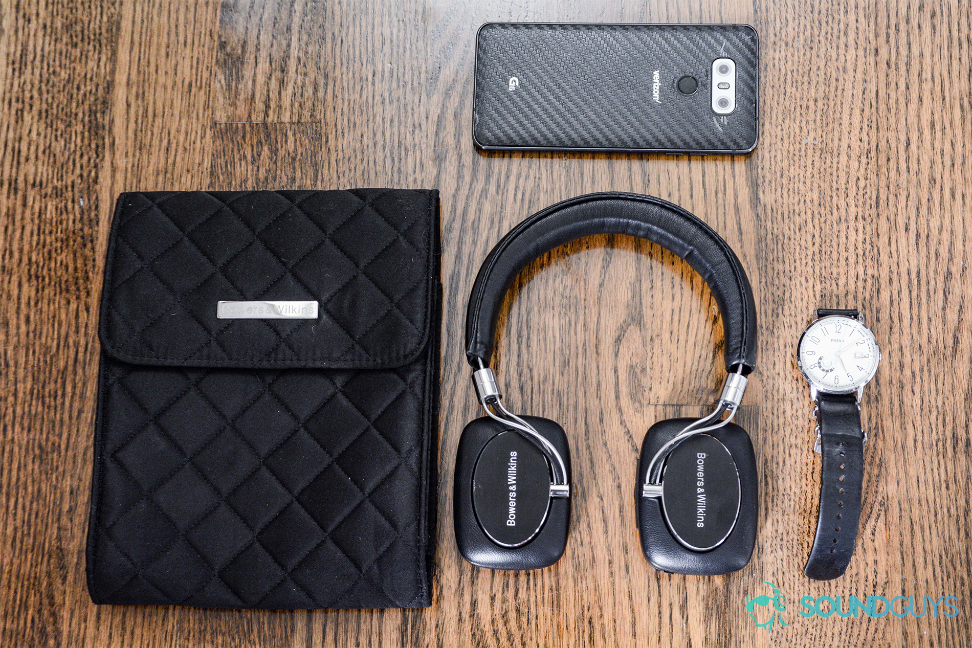 Bowers and Wilkins P5 Wireless: The headphones and quilted pouch on a wooden floor. A black watch and LG G6 appear too.