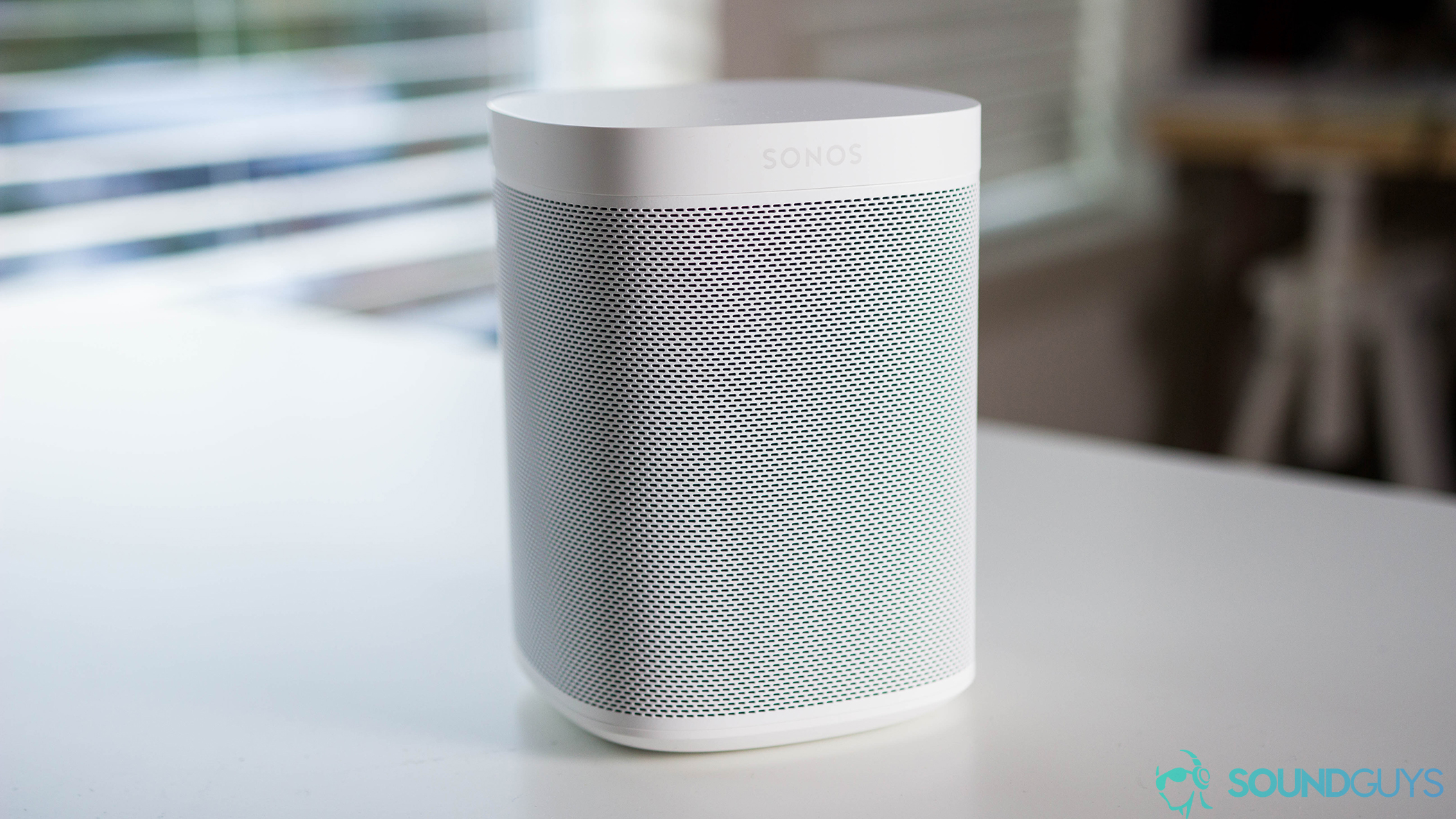 Picture of the Sonos One in white on a white surface with windows blinds in the background.