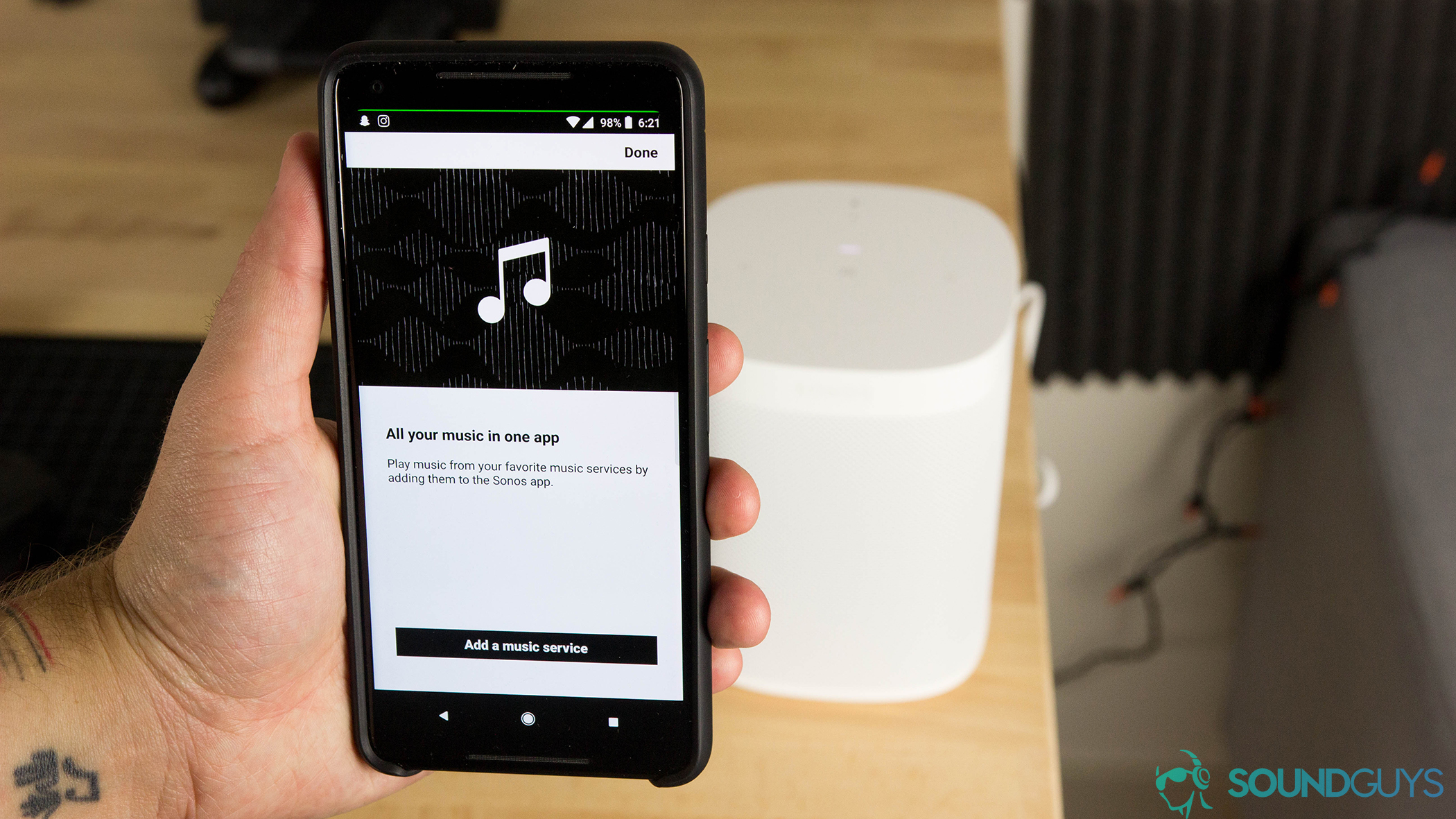 Image of a man holding an iPhone in front of a Sonos One (Gen 1) Google Assistant speaker (Alexa-supported) in white on a wood background.