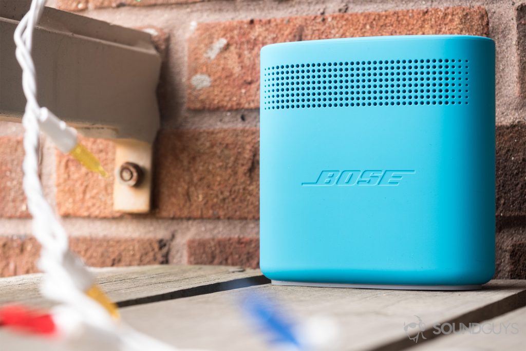 Dual-opposing, passive radiators are housed inside the Bose SoundLink Color II speaker. This minimizes surface vibrations while maximizing volume output. Pictured: The back of the Bose SoundLink Color II speaker, which has dotted openings on top-forth of the panel and the Bose logo emblazoned in the middle. It is ona wood balcony with decorative lights and a brick backdrop.