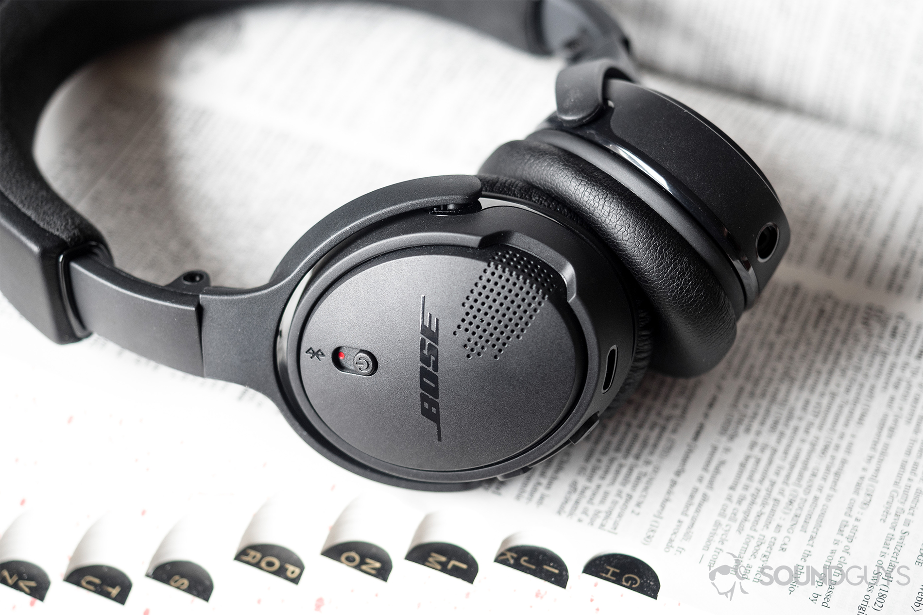 Made from a black plastic, the Bose On-Ear Wireless look classy and timeless, but their attenuated build may be cause for premature breakage. Pictured: The Bose On-Ear Wireless headphones with the right ear cup in focus, showing off the power switch and dual microphones. The headphones are laid flatly on an open dictionary.