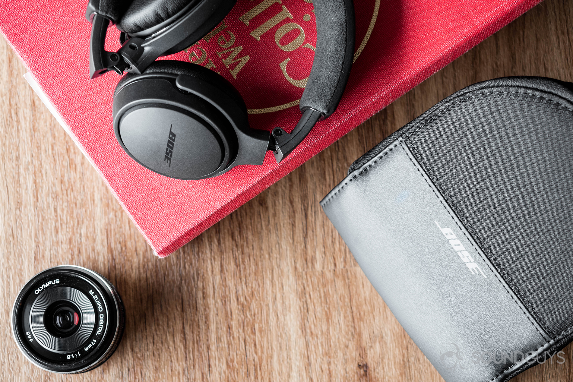 A photo of the Bose On-Ear Wireless headphones with the zippered carrying case and a lens in the corner. These are formed in a triangle-shaped position to balance out the frame, and the headphones are elevated on a red Webster's Dictionary.
