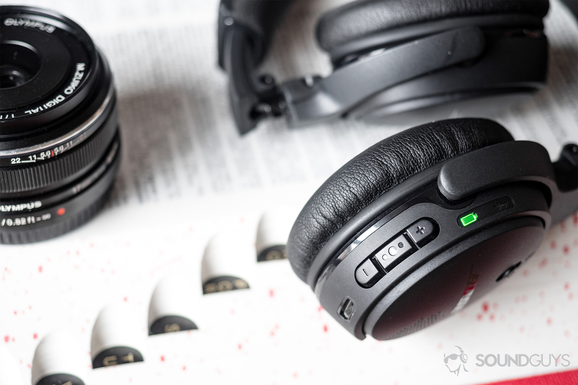 The battery life exceeds expectations by 30 minutes to a full hour, and the LED indicator informs listeners of the battery level. Pictured: The back of the right ear cup of the Bose On-Ear Wireless headphones with the battery LED lit up (green) to indicate that it is fully charged. The buttons (volume up, down, and multi-function) are also visible. The headphones are on an open dictionary with an Olympus M. Zuiko lens on the left side of the frame.