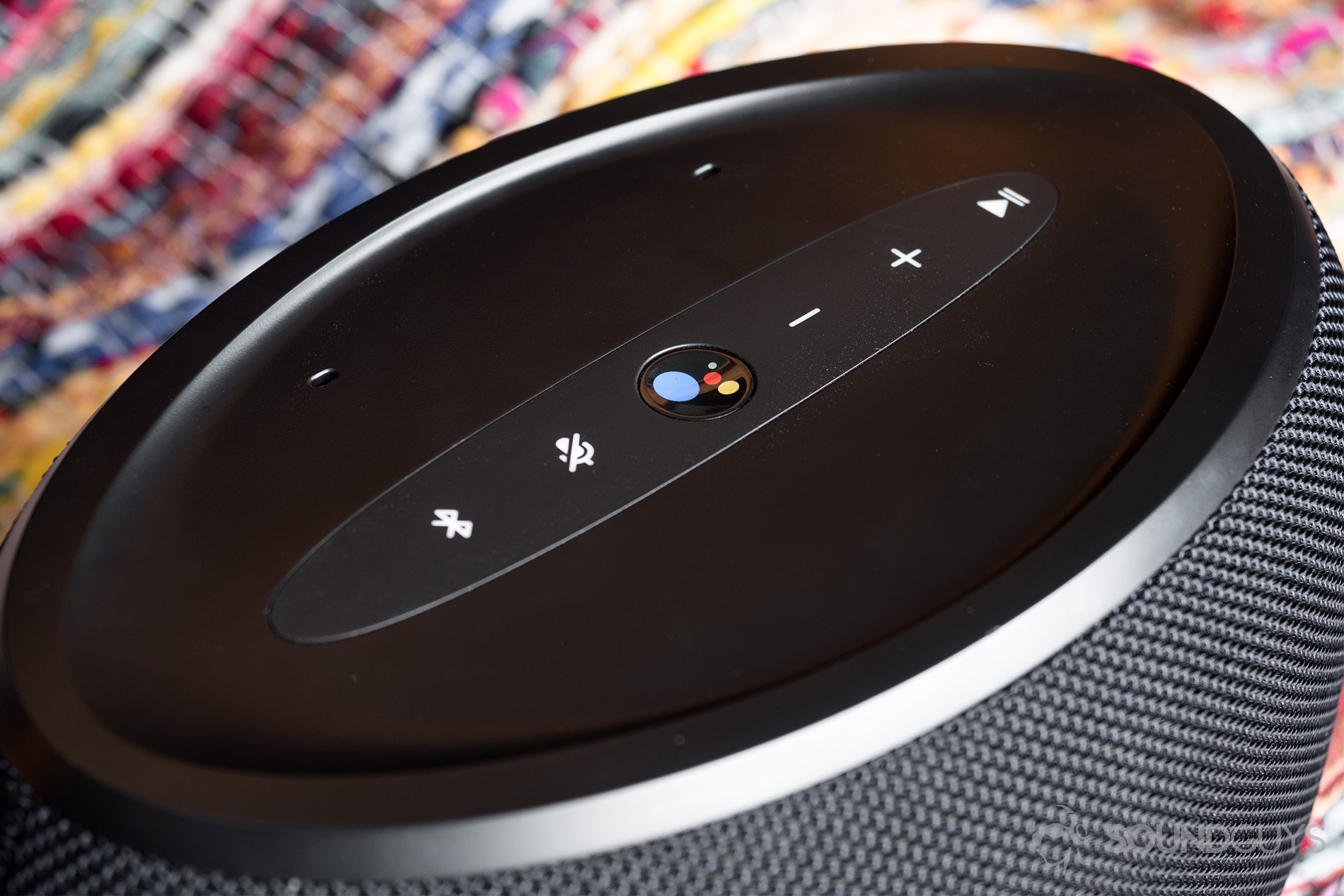 JBL Link 300 Google Assistant-integrated smart speaker: The speaker is viewed from a downward angle and runs diagonally across the screen. Featured are the controls on the top panel. In the centere is a colorized, Google Assistant button.
