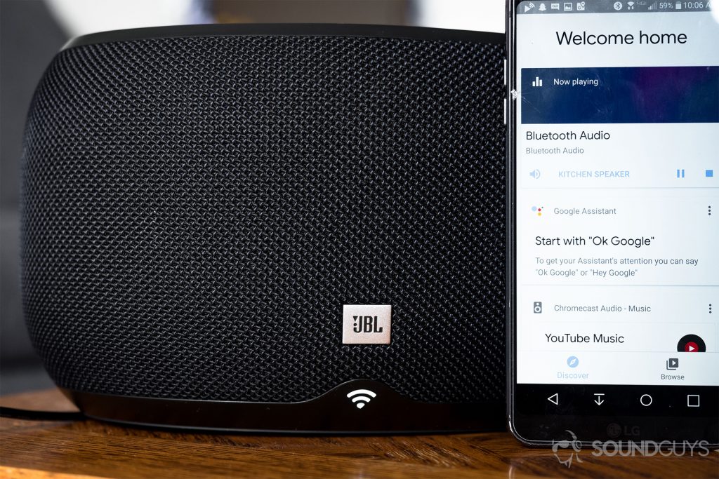 JBL Link 300 Google Assistant-integrated smart speaker: A straight-on shot of the black speaker wih the WiFI LED illuminated. On the right side is an LG G6 with the Google Home app pulled up.