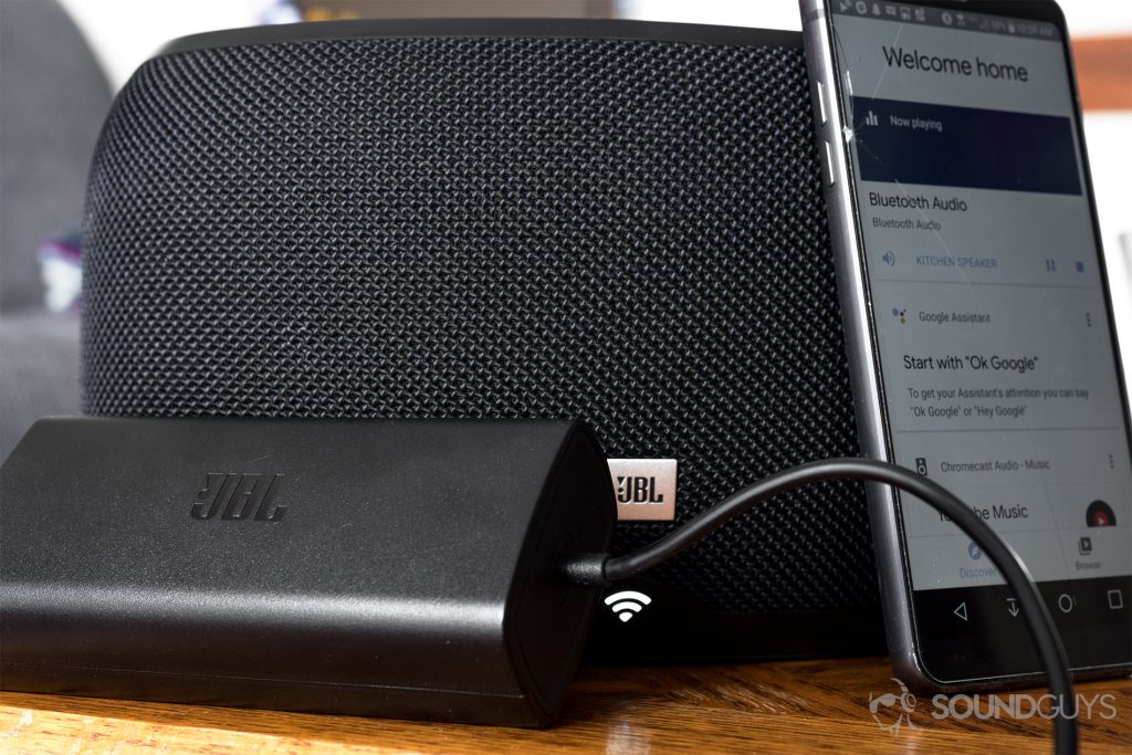 JBL Link 300 Google Assistant-integrated smart speaker: The included, JBL-branded power brick (black) in front of the speaker (black), and an angled LG G6 with the Google Home app active.