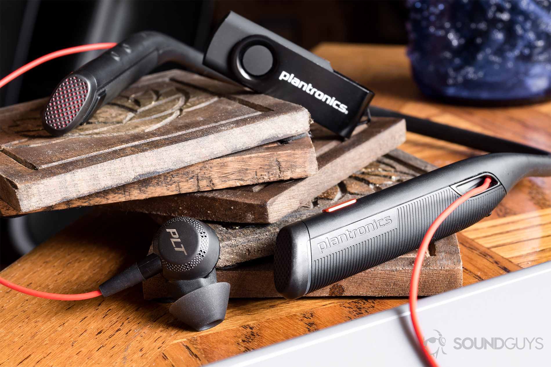 A basic, black paint job with orange accents keeps a low profile for the Plantronics Voyager 6200 UC headset. It's well-designed but could benefit from retractable earbuds. Pictured: The Plantronics headset on stacked, wood coasters on a wood table. The earbuds themselves are out of frame but the neckband is surrounding a Plantronics-branded flash drive that sits atop the coasters.