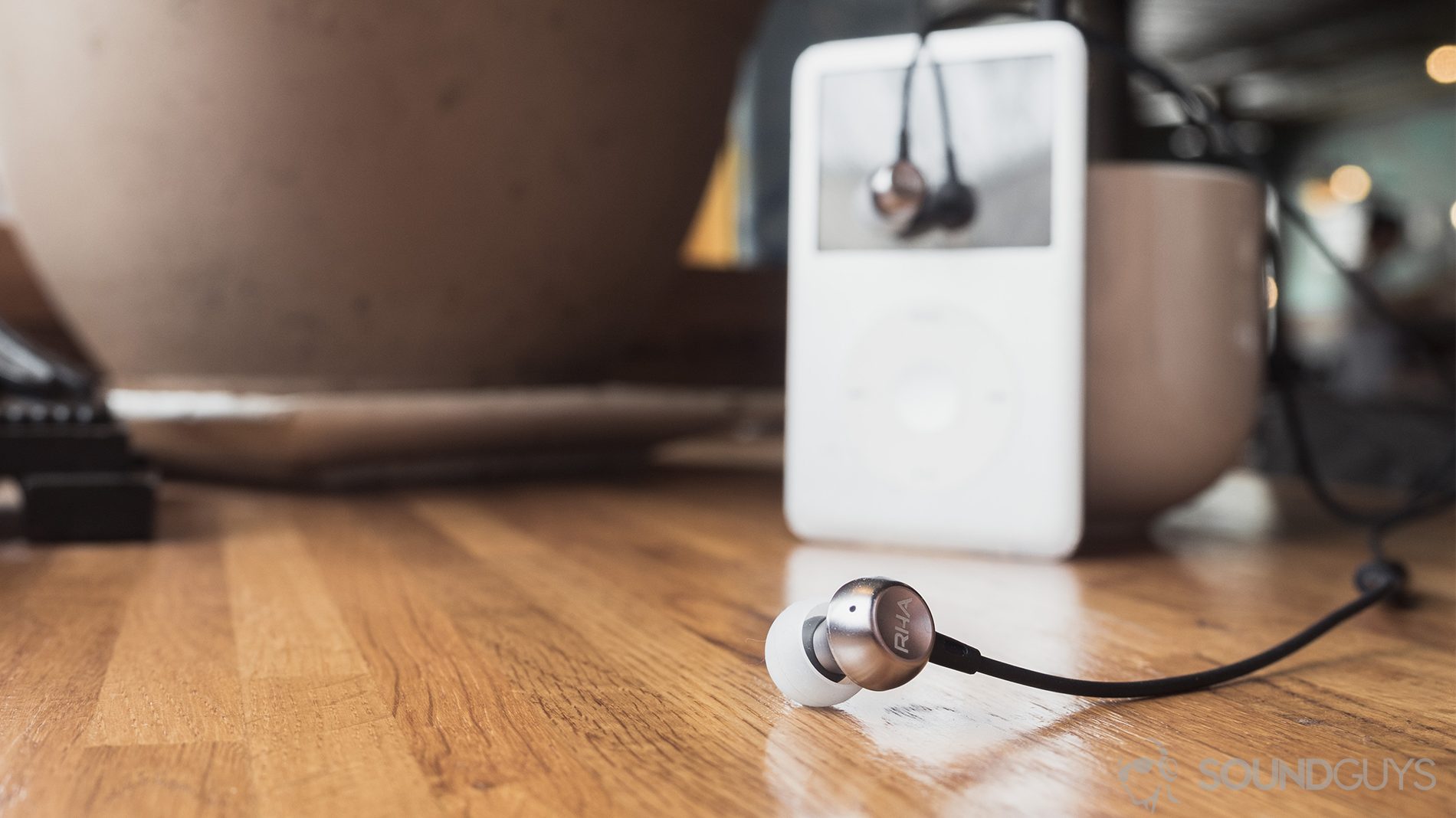 RHA MA390 review: The earbuds on a wooden surface. The background - an iPod classic stands against a large, white mug. The right earbud dangles over the screen of the iPod.