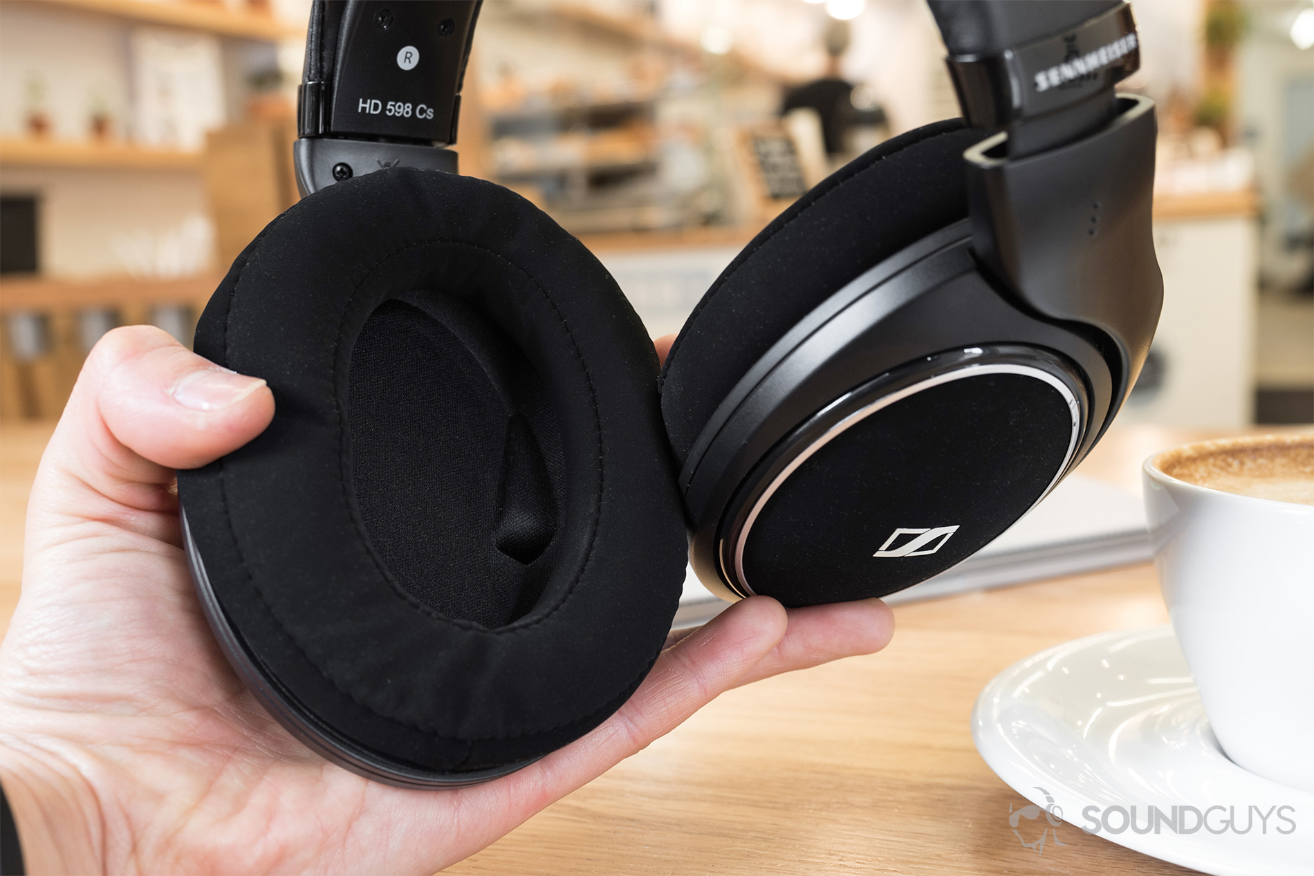 Sennheiser HD 598 CS review: The headphones in hand with the ear cups showing. Background: coffee shop with wood furnishing.
