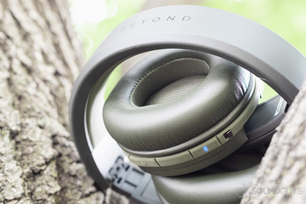 Focal Listen Wireless review: An underside view of the headphones (olive) which are folded up and placed beneath two thick branches of a tree. The Bluetooth LED (blue) is clearly visibile and active.