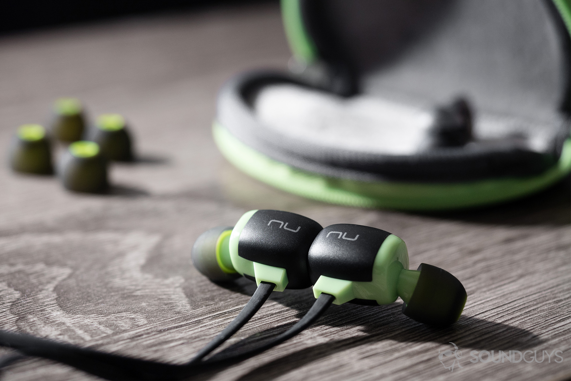 A picture of the Optoma NuForce BeSport4 wireless workout earbuds in green and grey with the carrying case and spare ear tips in the background.