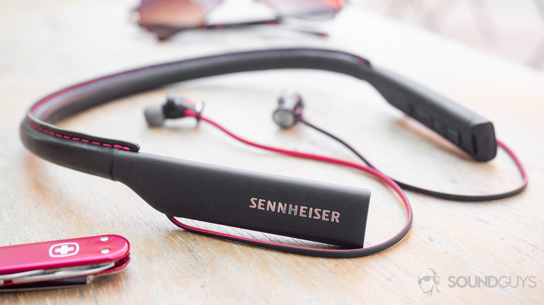 Sennheiser HD1 In-Ear Wireless earbuds on a wood table with sunglasses in the background and a Swiss Army knife in the foreground.