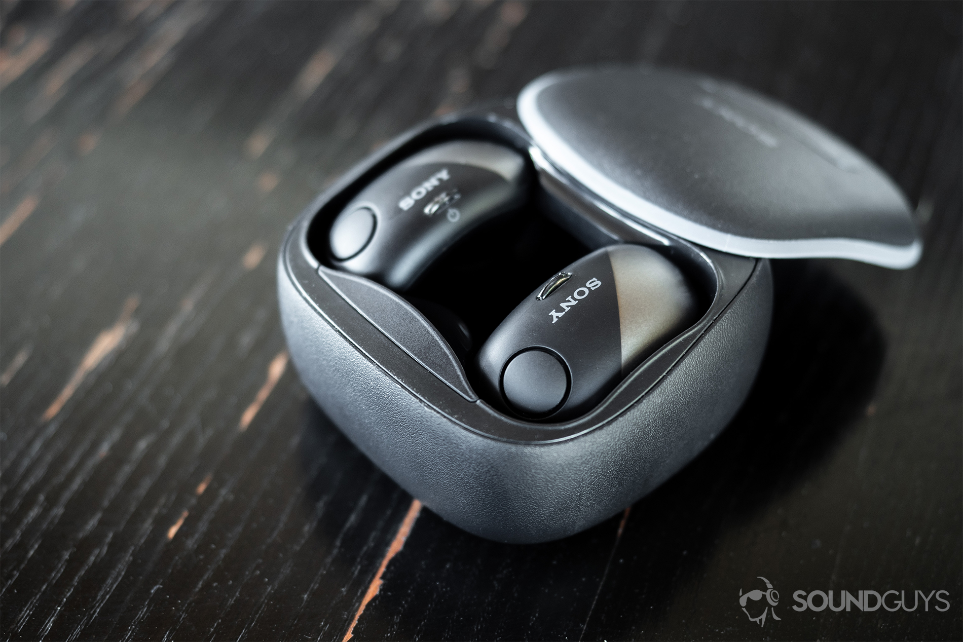 Sony_WF-700N-1 Sony WF-SP700N: A downward facing image of both true wireless earbuds docked in the charging case on a rustic, black wood surface.