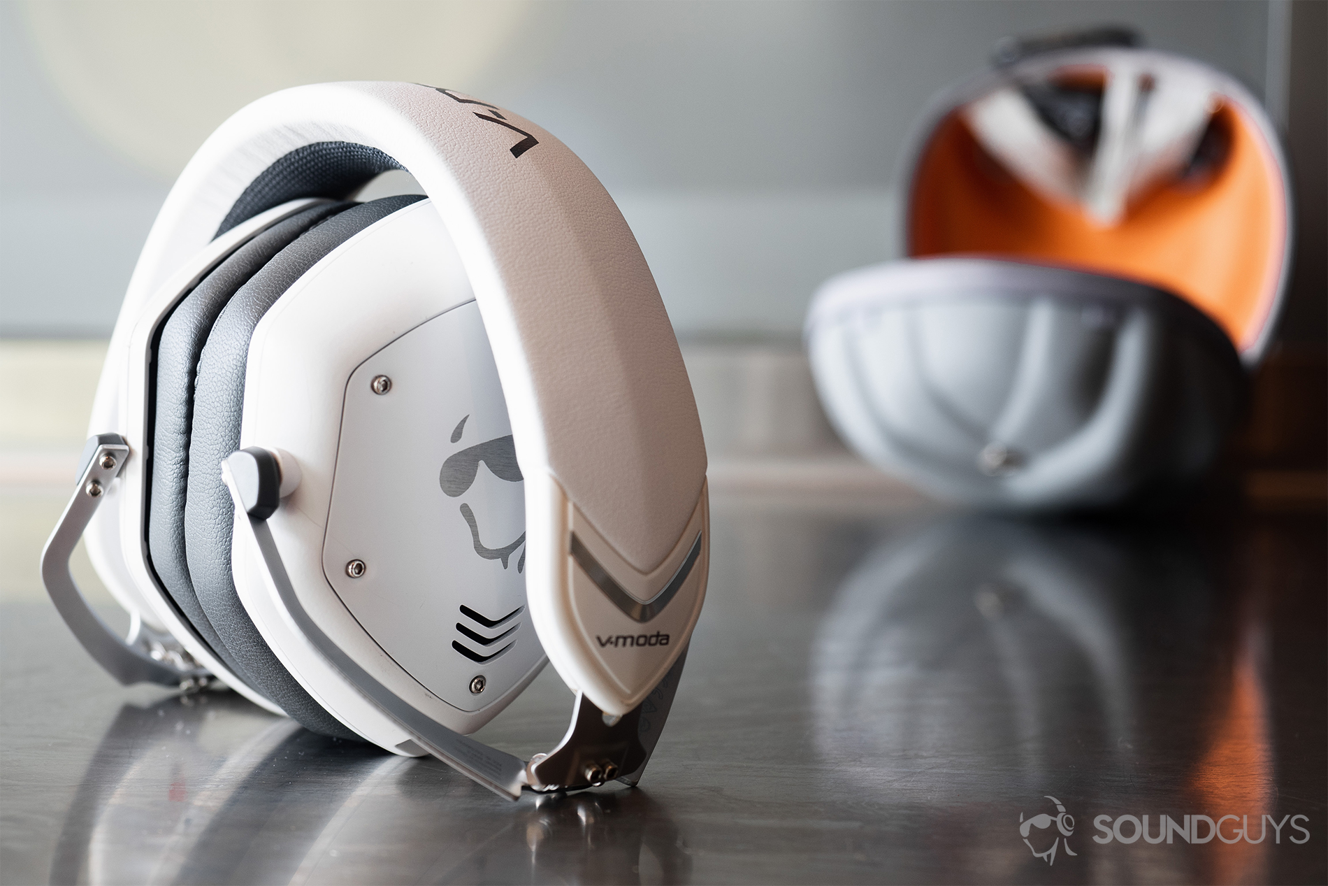 The V-Moda Crossfade 2 Codex headphones folded up but standing on a reflective surface wtih the clam shell Exoskeleton case in the background.