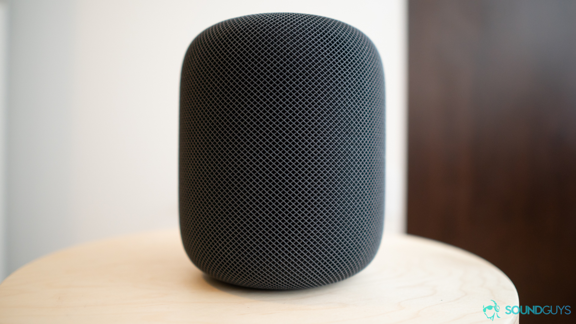 The black Apple Homepod on a wooden stool against a white wall. 