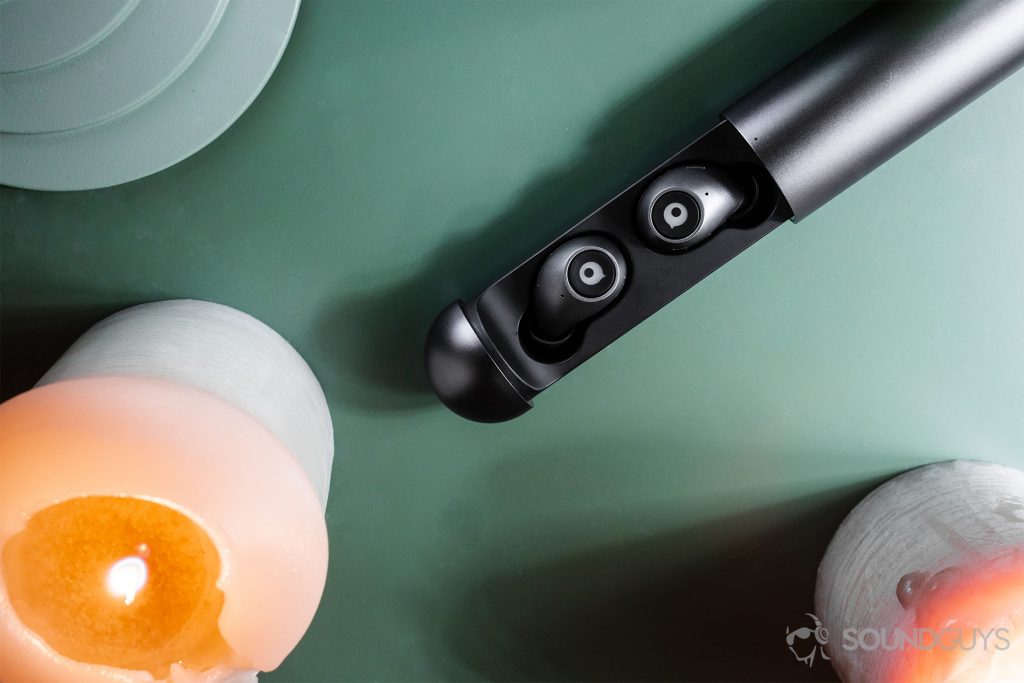 A top-down image of the earbuds in the included charging case which is unsheathed. There is a candle in the lower left and lower right corners. The surface is a mellow green.