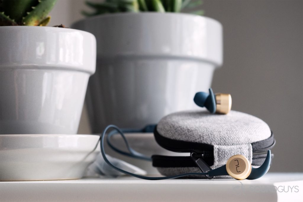 Optoma NuForce Be Live5: The earbuds on a white table with their included carring case. Potted succulents in the background; soft lighting.
