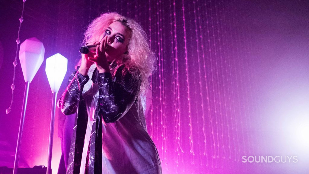 A woman sings into a microphone on a magenta lit stage.