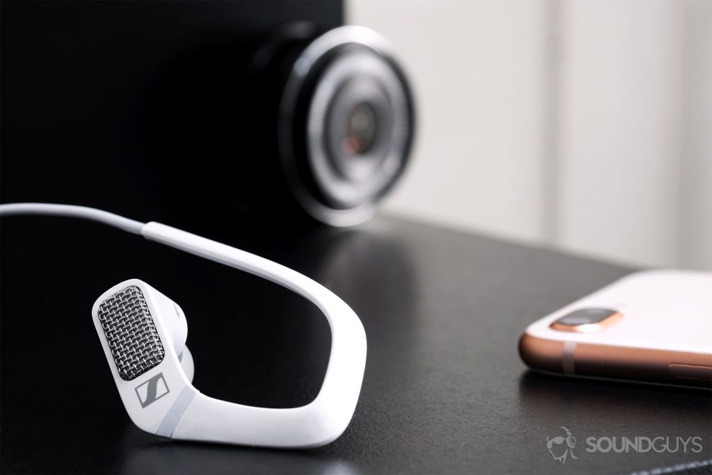 Sennheiser Ambeo Headset: The right earbud in the foreground (left) with an iPhone 8 Plus on the poking in from the right side of the image. An olympus lens is in the background.