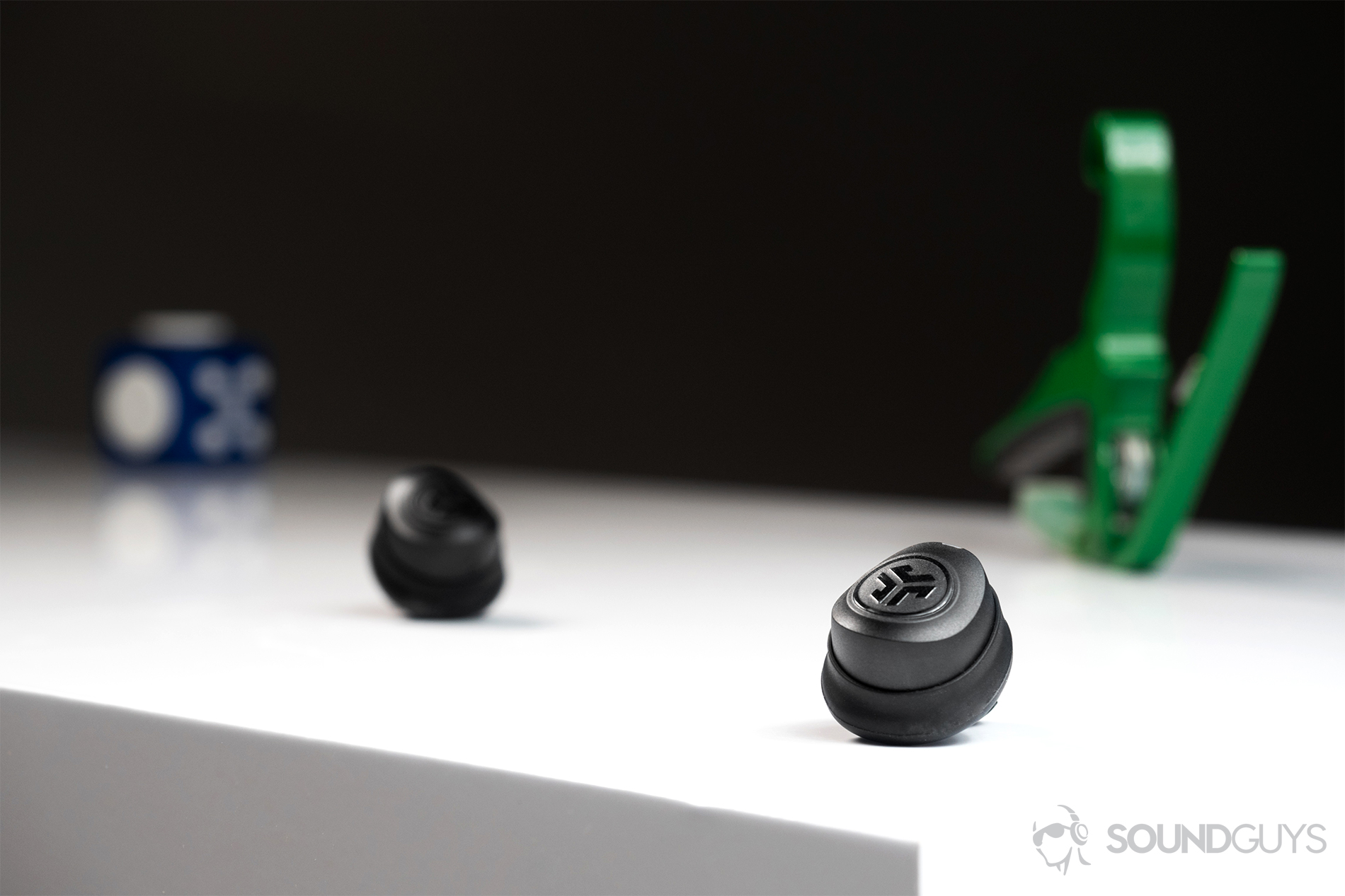 A picture of the JLab JBuds Air true wireless workout earbuds (black) on a white table with a guitar capo in the background.