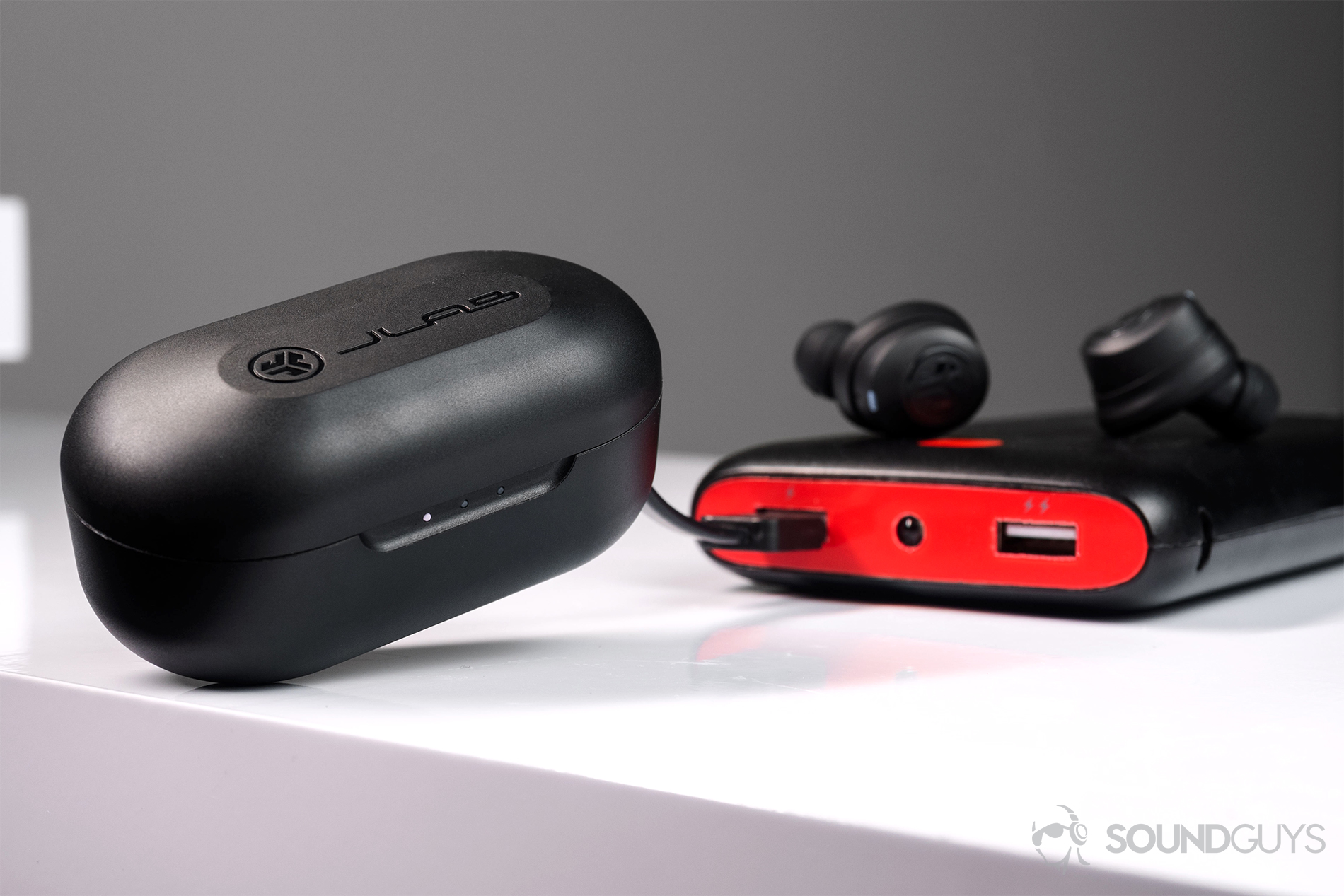 JLab JBuds Air true wireless charging case connected to a portable battery pack with the earbuds resting on the pack.