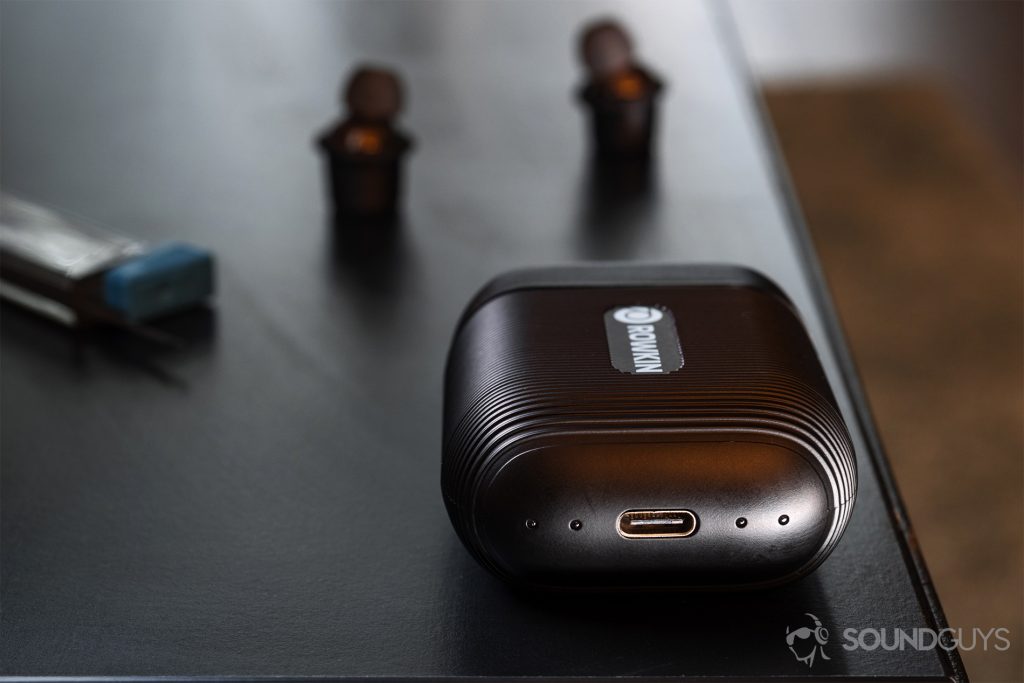 Rowkin Ascent Micro: The charging case with the USB-C port in focus and the earbuds in teh background. All objects rest on a black table.