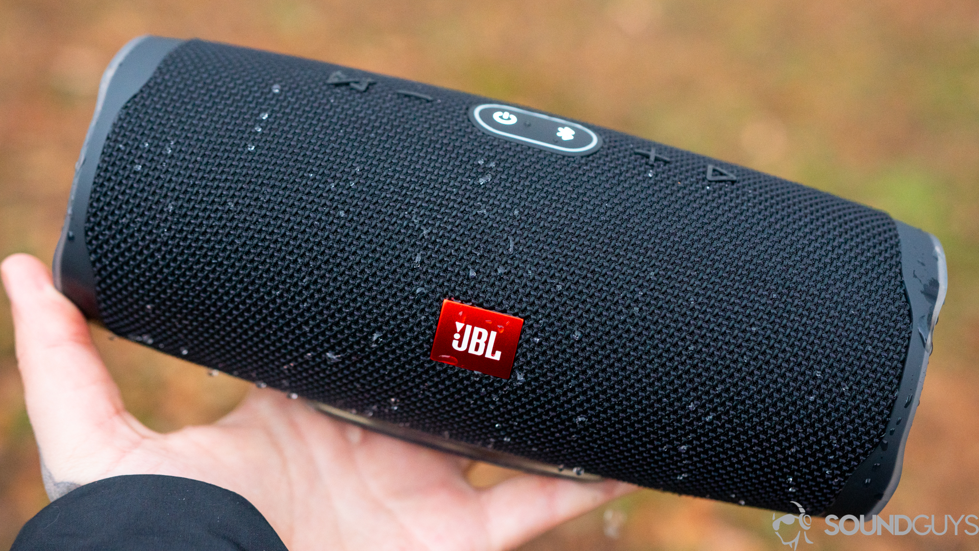 Man holding a wet all-black JBL Charge 4 in hand with water droplets covering it. 