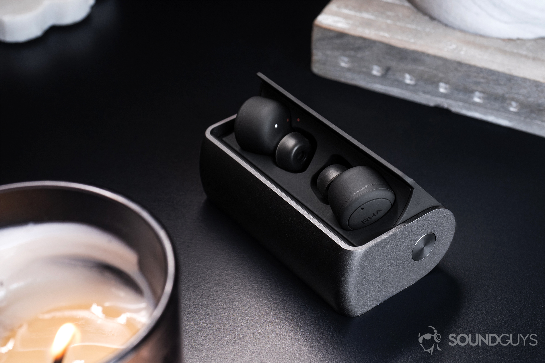 RHA TrueConnect true wireless earbuds in the open charging case which rests on a black table. A candle sits in the bottom left corner of the image.