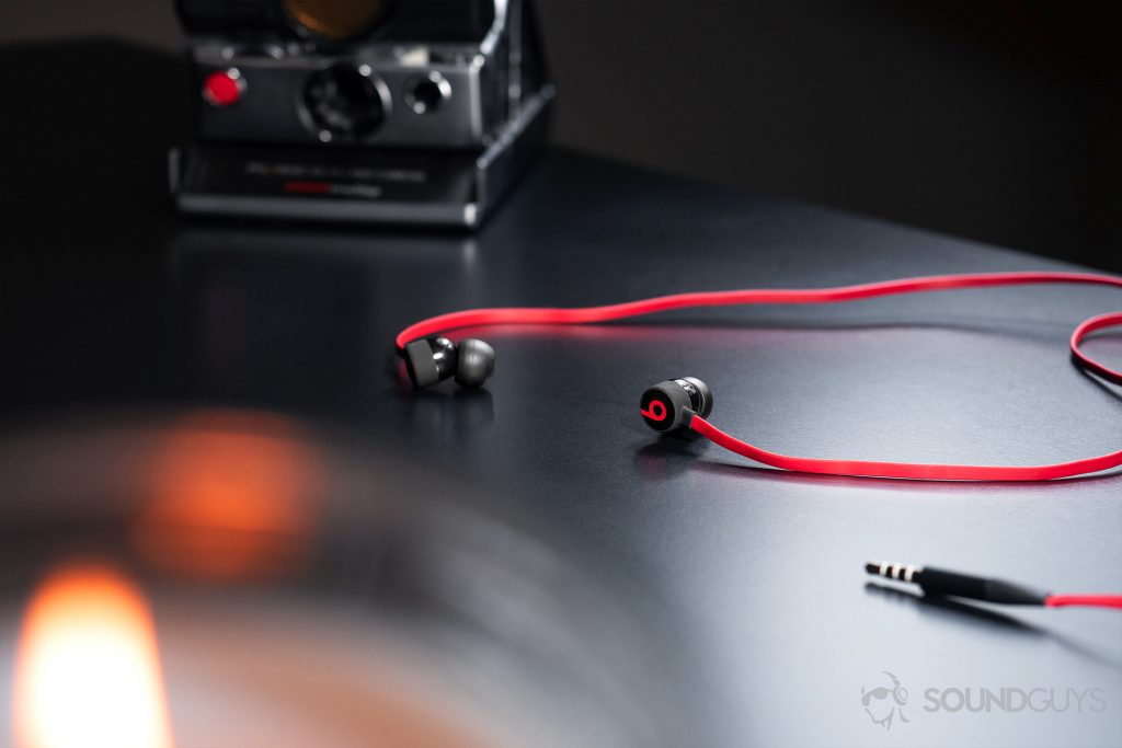 Beats urBeats3: The earbuds resting on a black table with a vintage camera in the background and blurred out part of a candle in the foreground.
