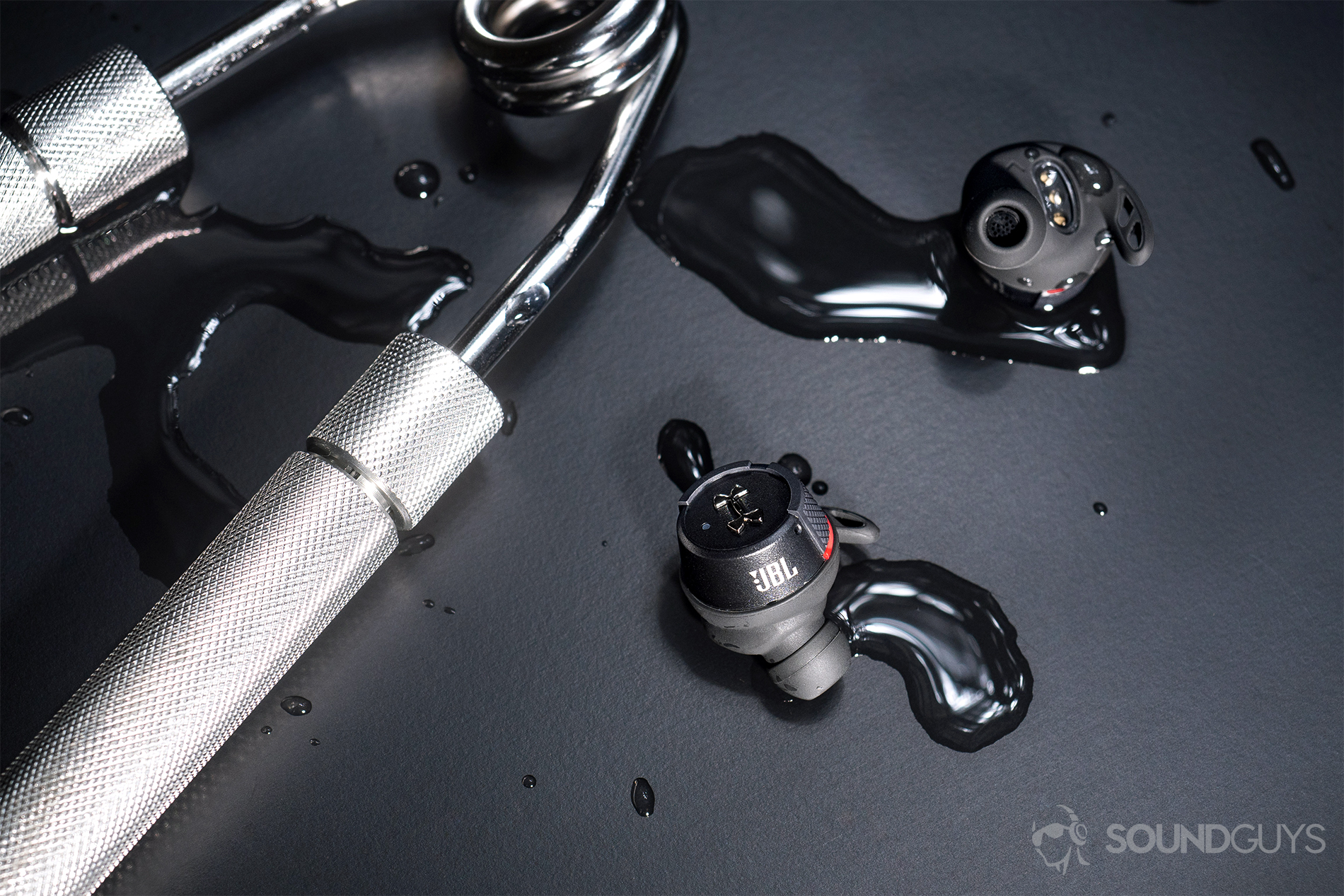 Under Armour_True Wireless Flash by JBL-6 A picture of the Under Armour True Wireless Flash JBL: Top-down image of the earbuds next to a grip strength piece of exercise equipment. There's water on the table and earbuds.