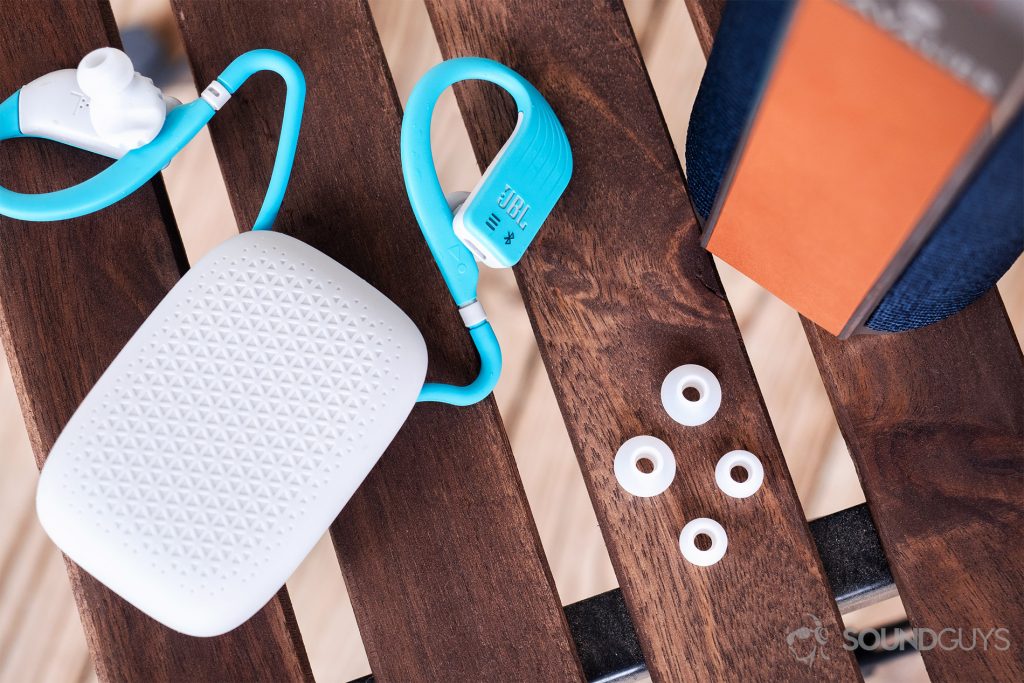 JBL Endurance Dive: Top-down image of the headphones coming out of the silicone pouch with the included ear tips to the side. A Cavalier speaker is in the top right corner of the image and all objects are on slatted wood.