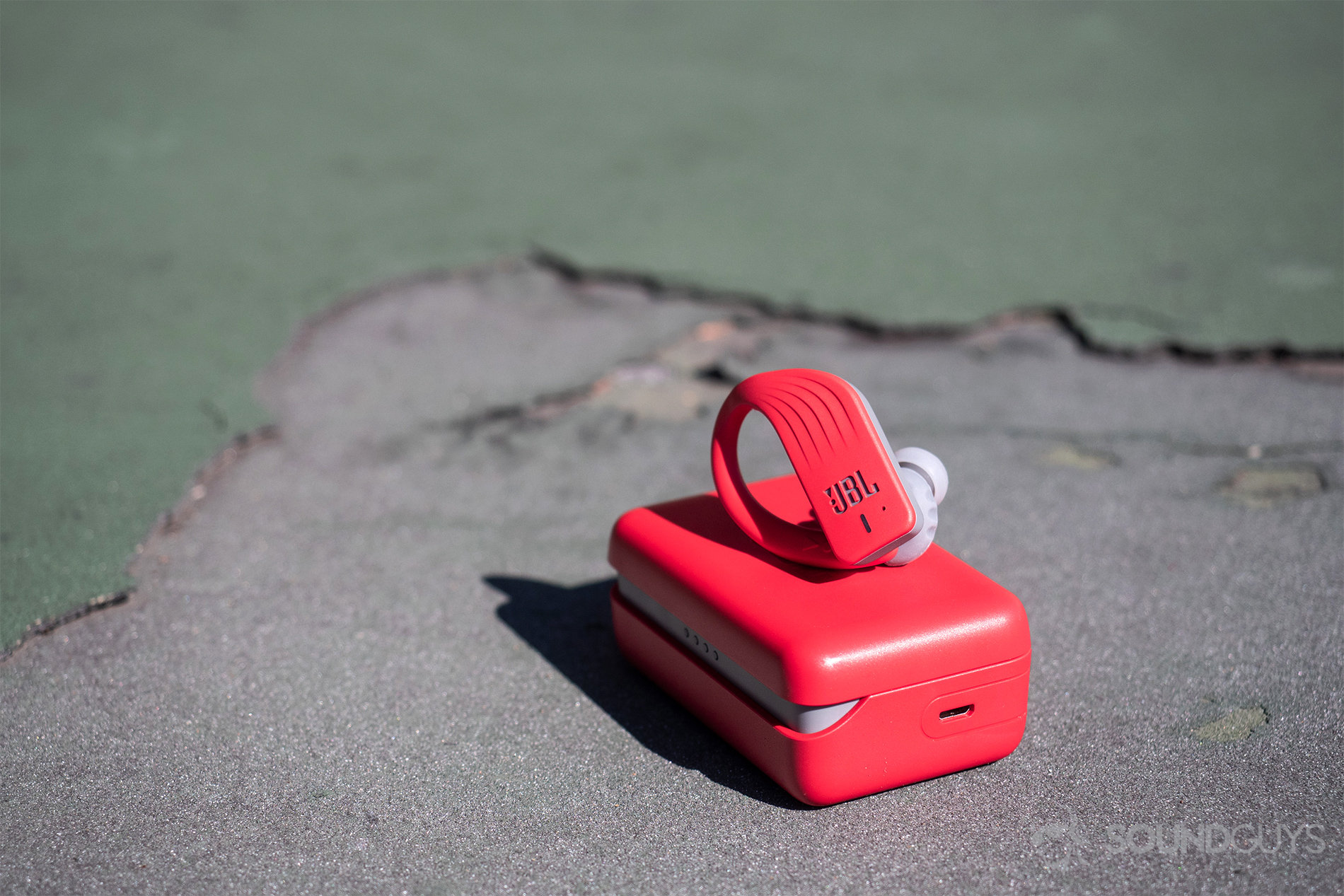 JBL Endurance Peak: The right earbud resting on the case, both of which are angled toward the camera and resting on a tennis court.