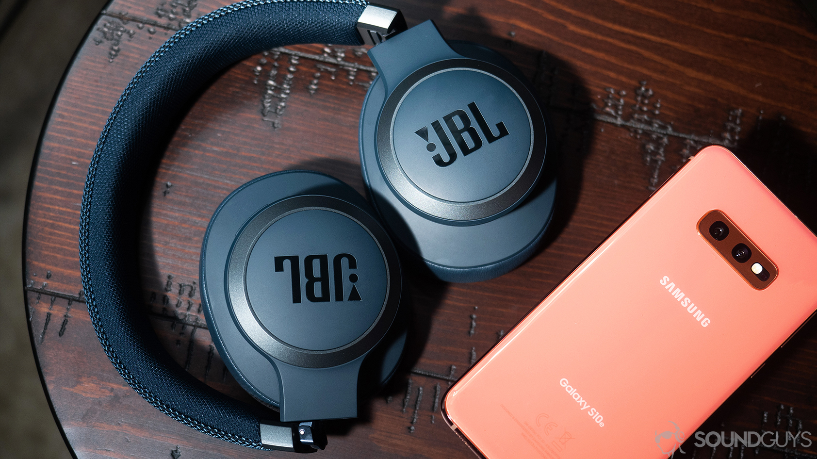 A picture of the JBL Live 650BTNC noise canceling headphones folded up and lying flat on a wooden surface by a Samsung Galaxy S10e smartphone.