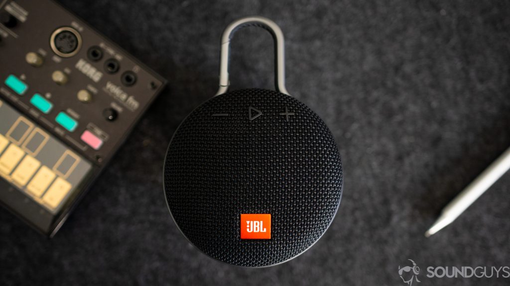 Pictured is the JBL Clip 4 against a gray background with a synthesize and pen. 