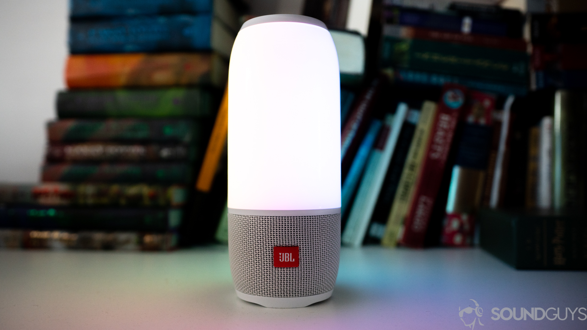 The white JBL Pulse 3 in front of colorful books on a white desk.