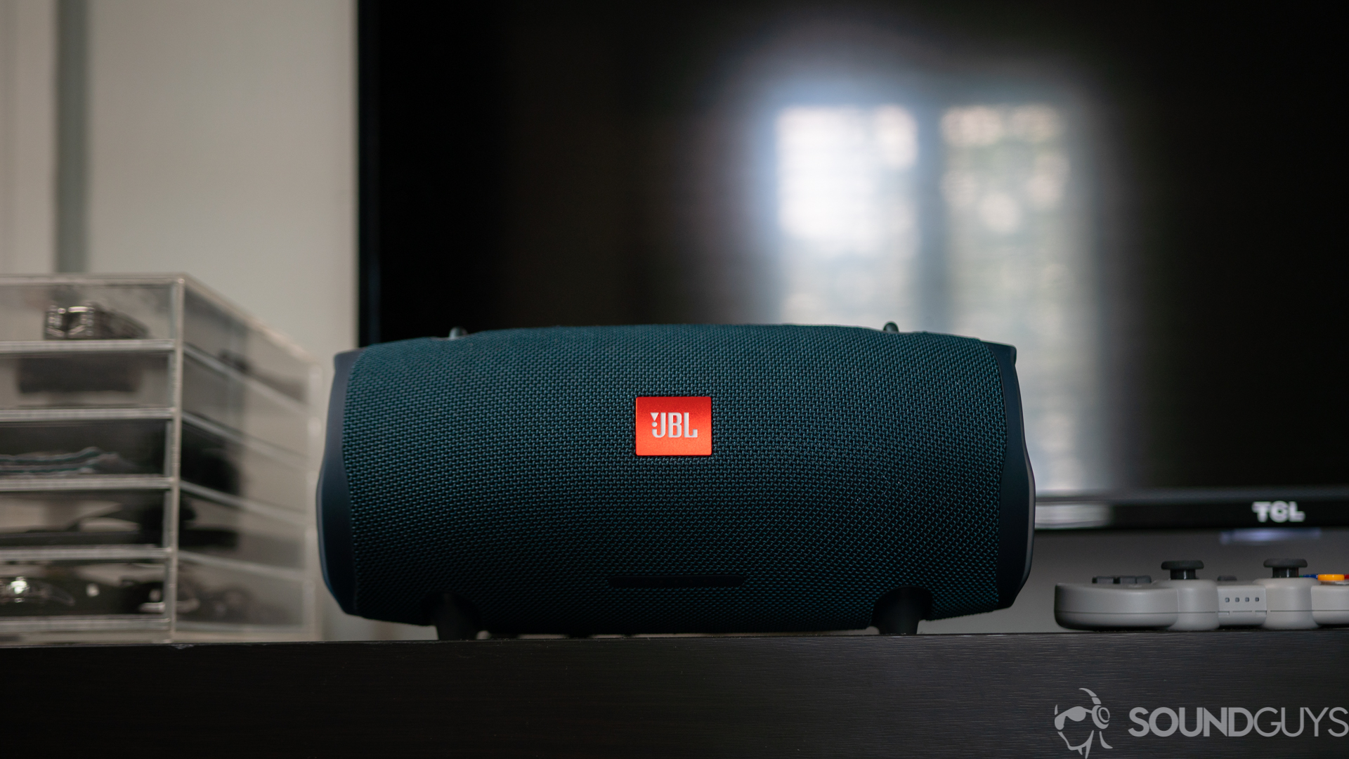 Shot of the JBL Xtreme 2 from the front on top of a black table with a TV in the background.
