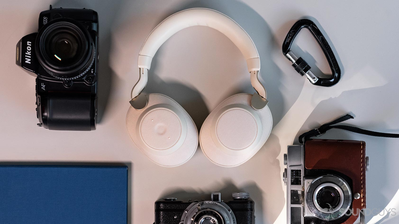 The Jabra Elite 85h headphones folded flat on a table and surrounded by vintage cameras, a blue notebook, and a black carabiner.