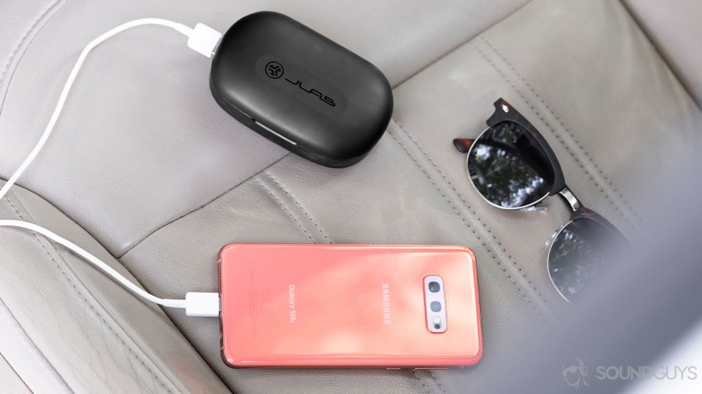 The JLab JBuds Epic Air charging case charging a Samsung Galaxy S10e in Flamingo Pink. A pair of sunglasses are next to the items on a leather car seat.