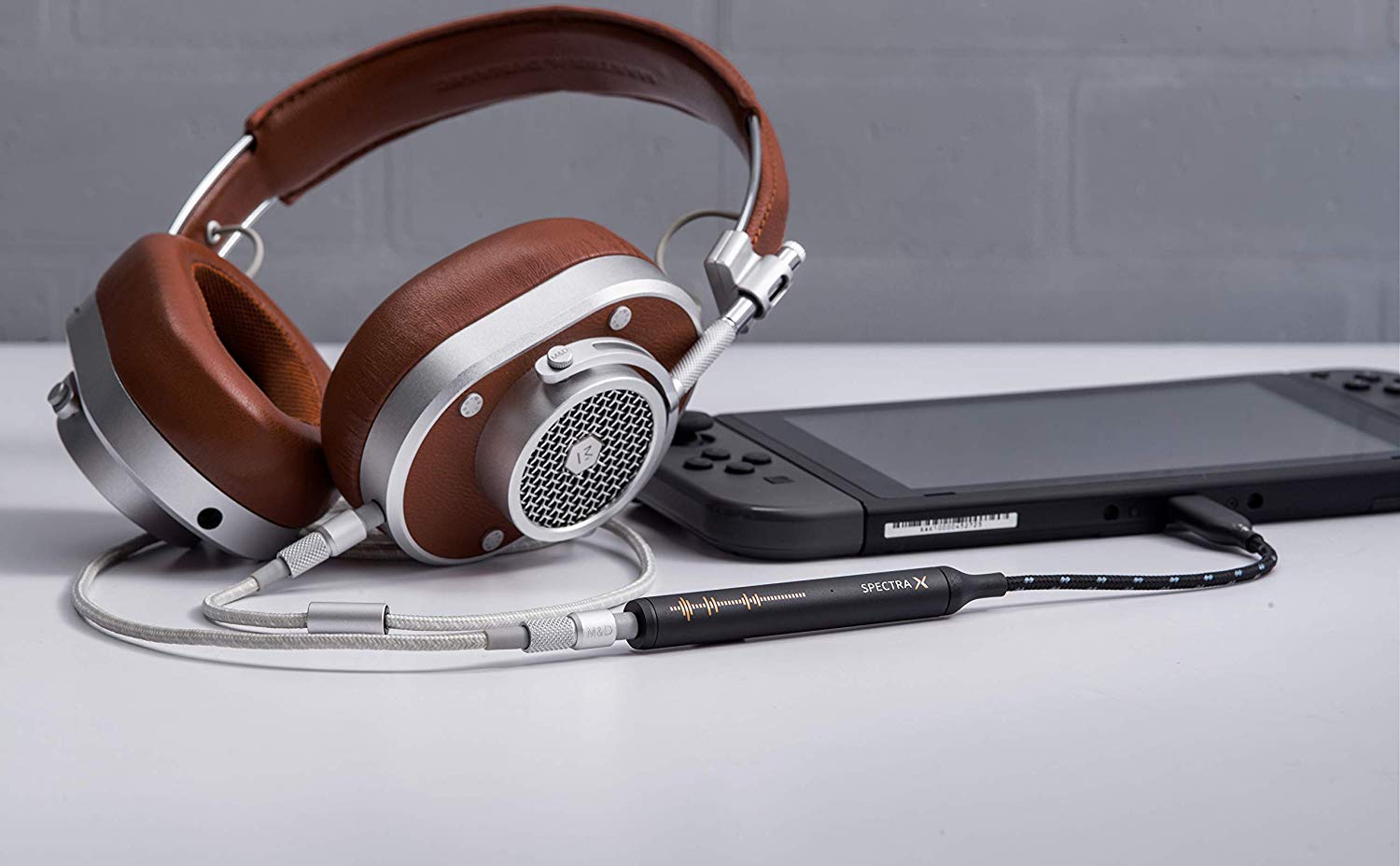 NextDrive Spectra X amplifier plugged into Master & Dynamic brown leather headphones. Also connected to a Nintendo Switch in black. All items are on a white table.