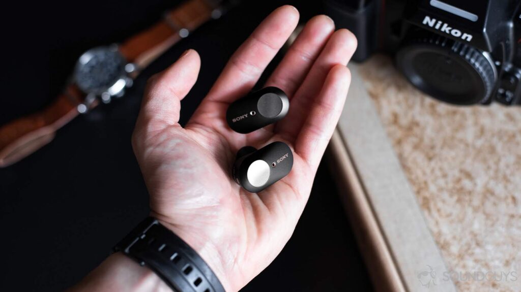 A photo of the Sony WF-1000XM3 earbuds in the palm of a left hand with a beige book and leather watch on a table in the background.