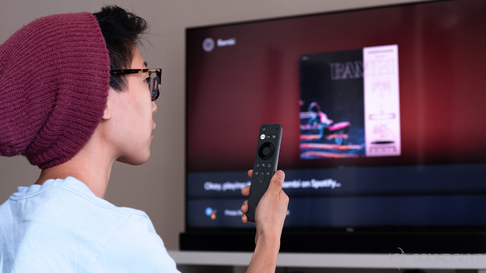 A woman holding the JBL Link Bar remote (a wireless JBL speaker) and speaking to Google Assistant to switch songs on Spotify.