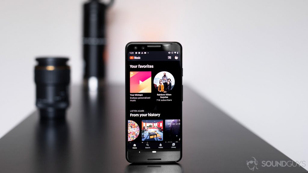 A picture of the Google Pixel 3 with the YouTube Music Premium app open to the home screen. The phone is standing vertically on a black table with a lens and water bottle in the background.