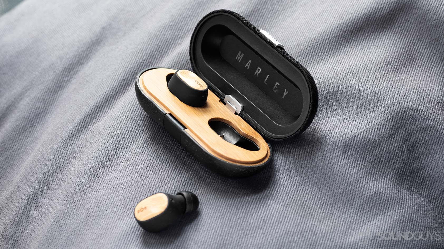The House of Marley Liberate Air earbuds. One is in the charging case, which is open, while the other is on the grey, cloth surface.