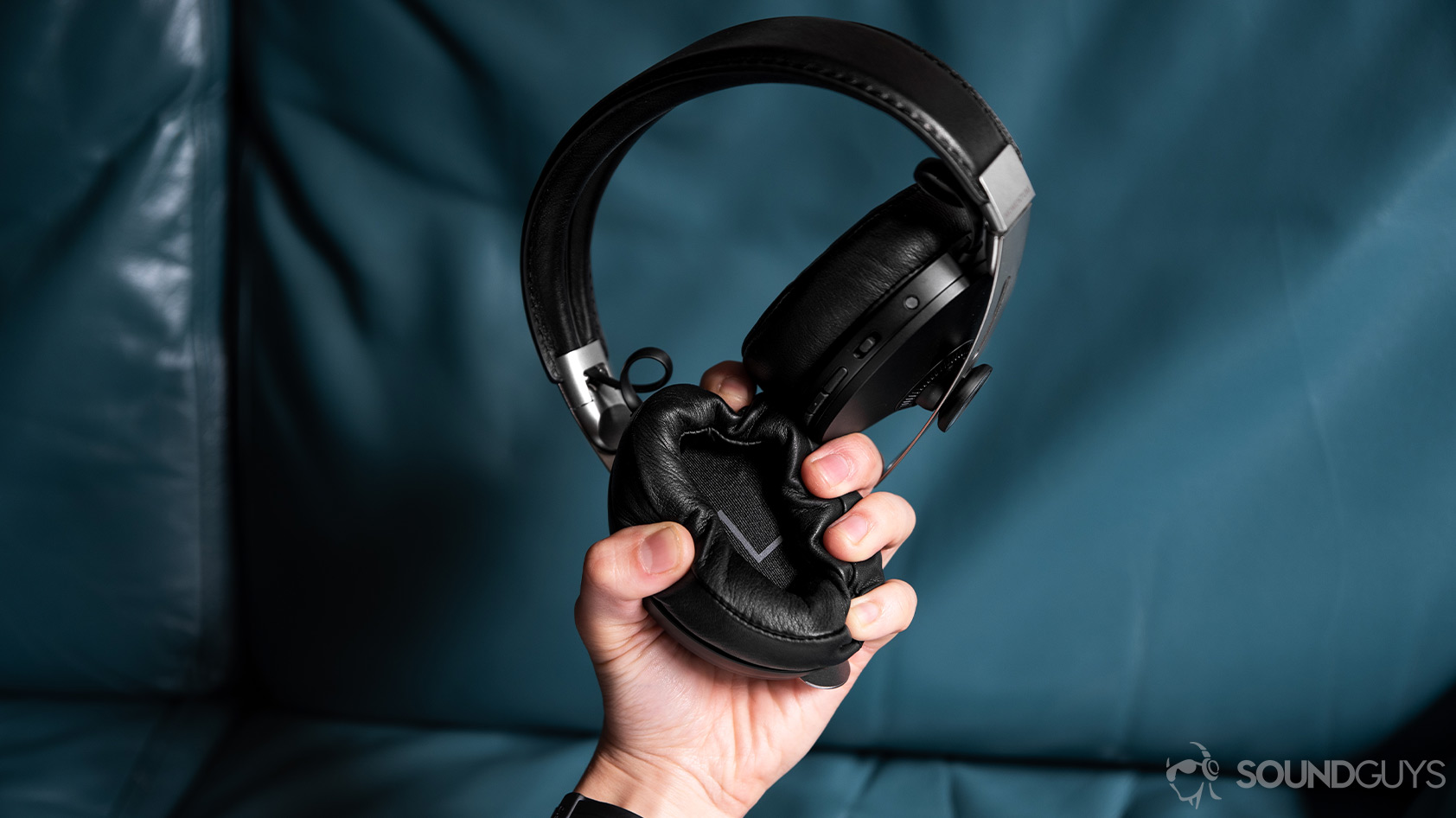 The Sennheiser Momentum Wireless 3 left ear pad begin crushed in the hand in front of a teal couch.