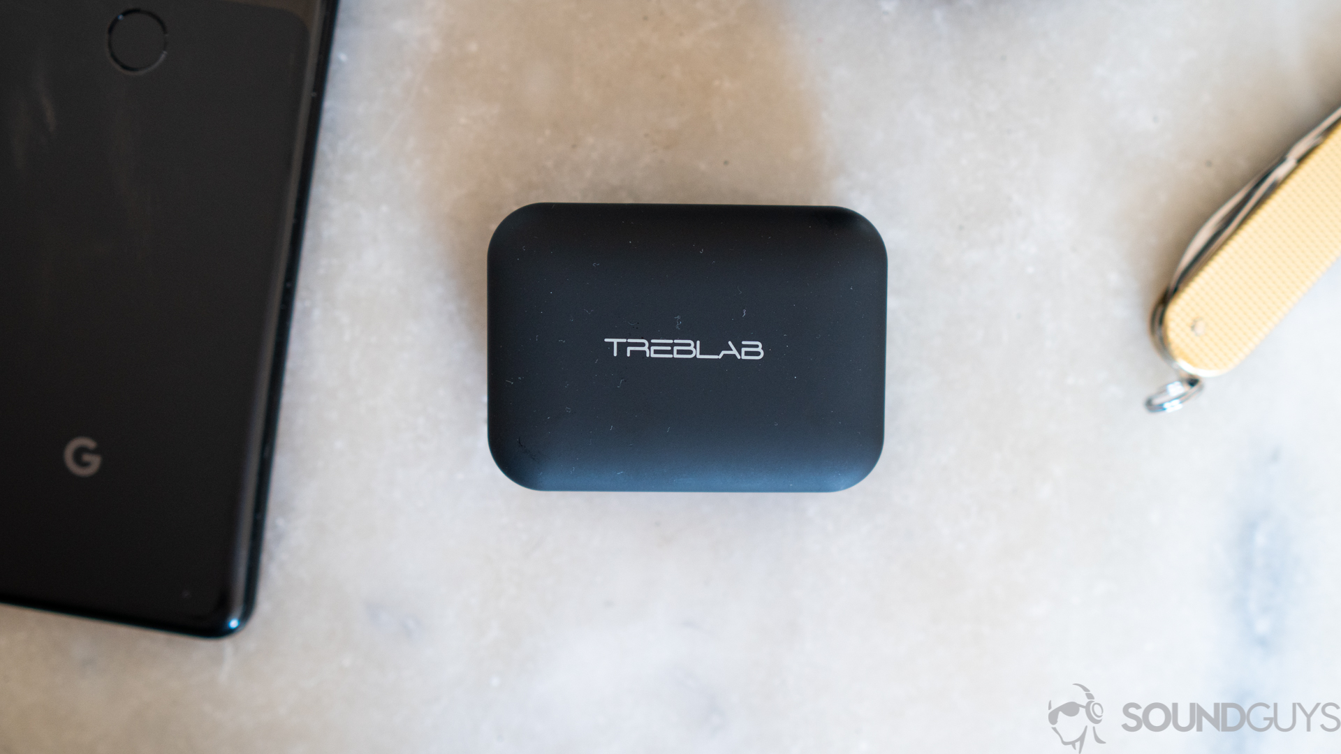 A photo of the Treblab X5 in its carrying case, resting atop a table, next to a Google Pixel phone and pocketknife.